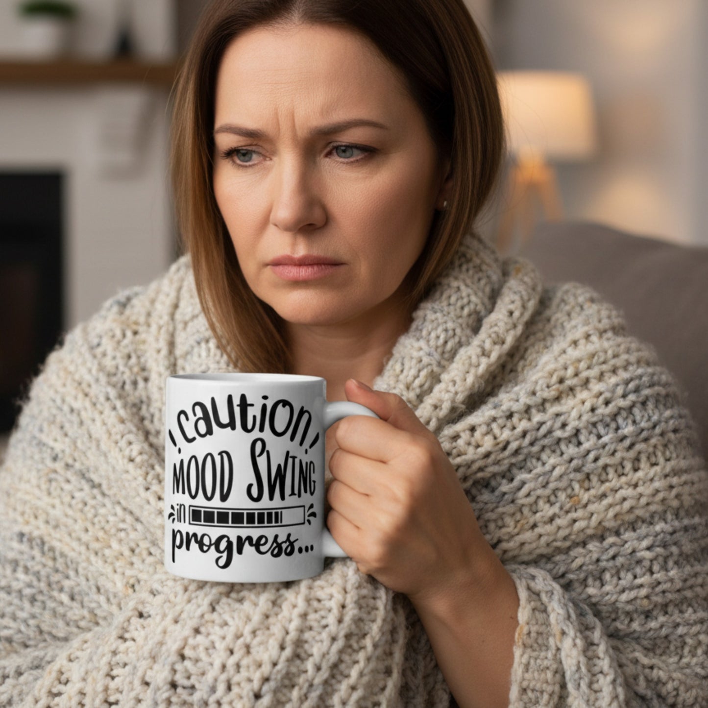Woman wrapped in a cosy knitted blanket holding a Caution Mood Swing In Progress funny coffee mug. Lifestyle mockup showing a humorous mood swing mug for coffee lovers, menopause warriors, and funny office gift ideas.