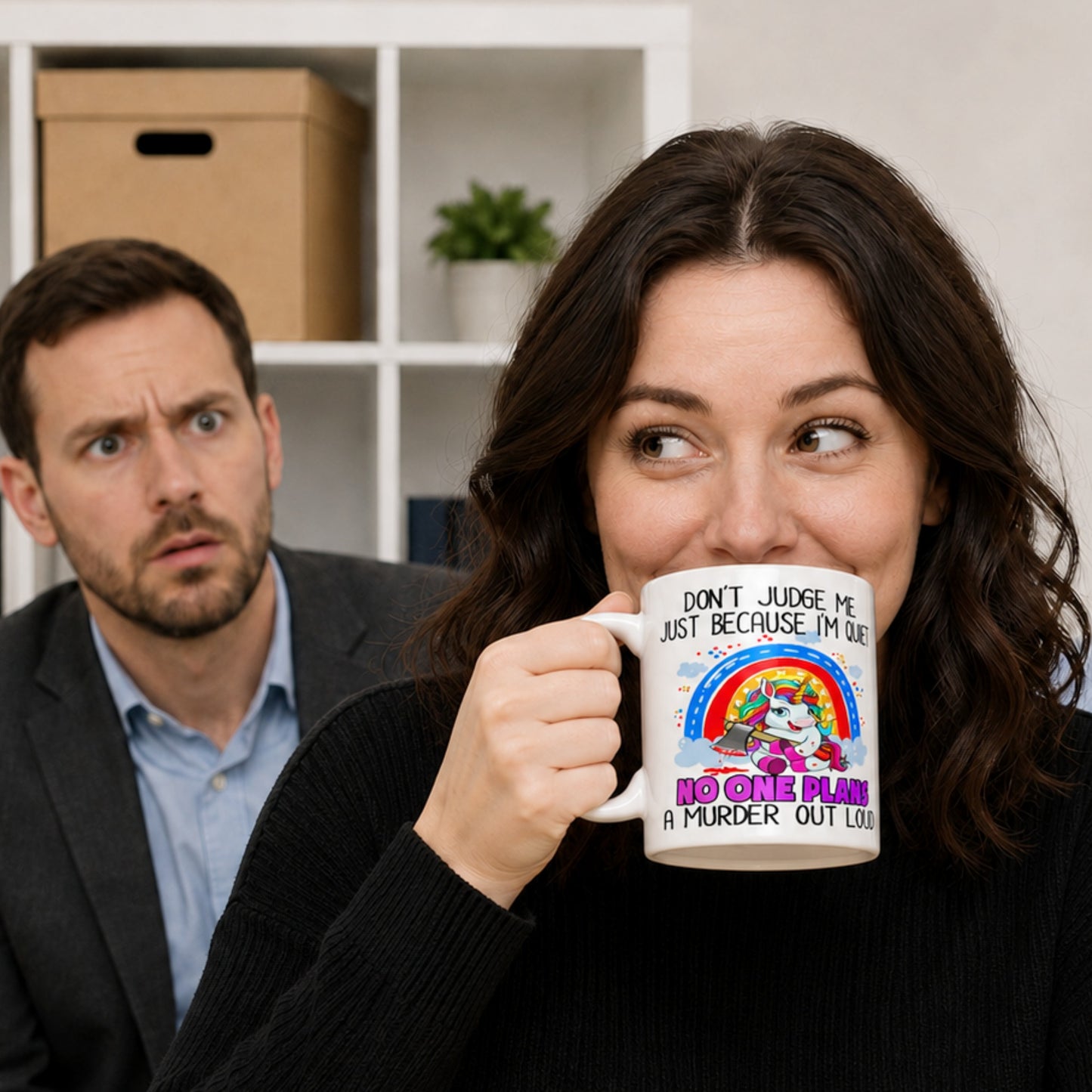 Woman smiling while drinking from a funny sarcastic unicorn coffee mug that reads "Don't Judge Me Because I'm Quiet, No One Plans A Murder Out Loud," while a shocked coworker looks on in the background. Humorous office lifestyle mockup featuring a novelty ceramic mug with dark humour.