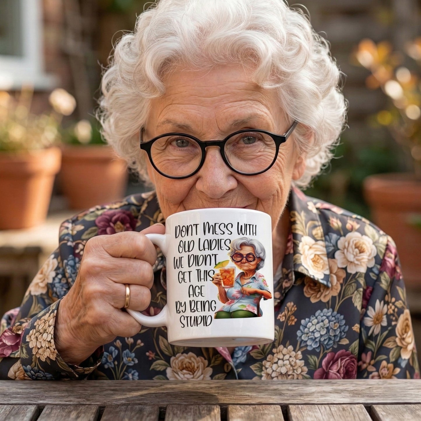 Smiling older woman enjoying a Don't Mess With Old Ladies funny grandma coffee mug while sitting outdoors, showcasing the sarcastic novelty mug in a natural lifestyle setting.