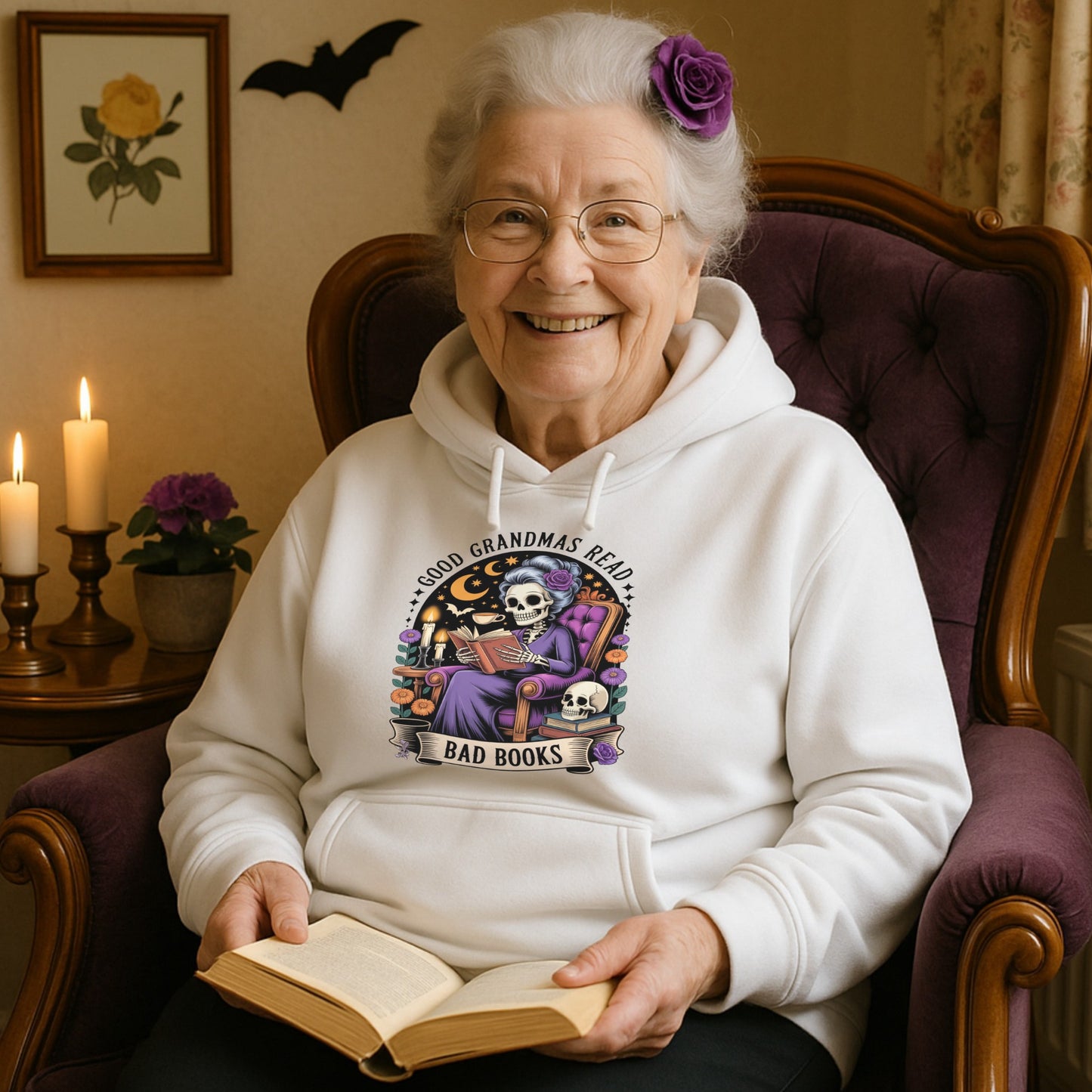Woman wearing a hoodie with a graphic design, with a spooky-cute skeleton grandma reading a “bad” book under moonlight – the perfect mix of bookish and gothic vibes. sitting in a chair holding a book.