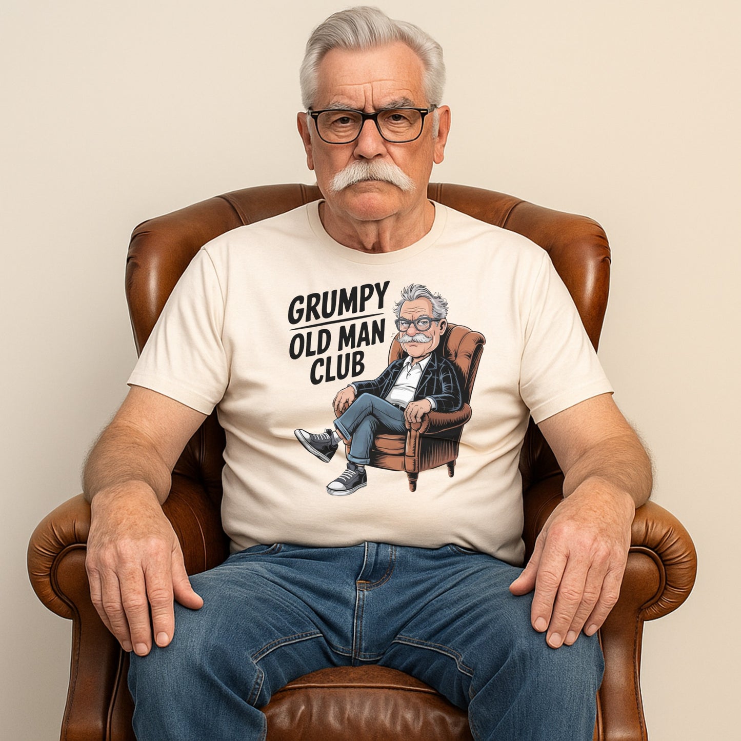 An older man wearing a "Grumpy Old Man Club" t-shirt with a cartoon of a grumpy man in an armchair, sitting seriously in a leather chair.