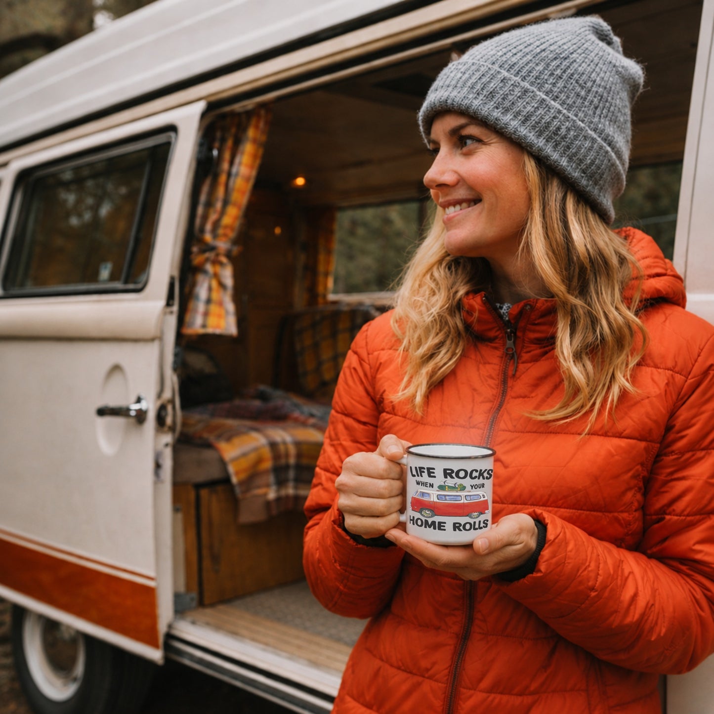 Lifestyle mockup of the Life Rocks When Your Home Rolls outdoor enamel mug held by a woman outside a vintage campervan, featuring a red camper van design during a cosy camping and van life adventure.