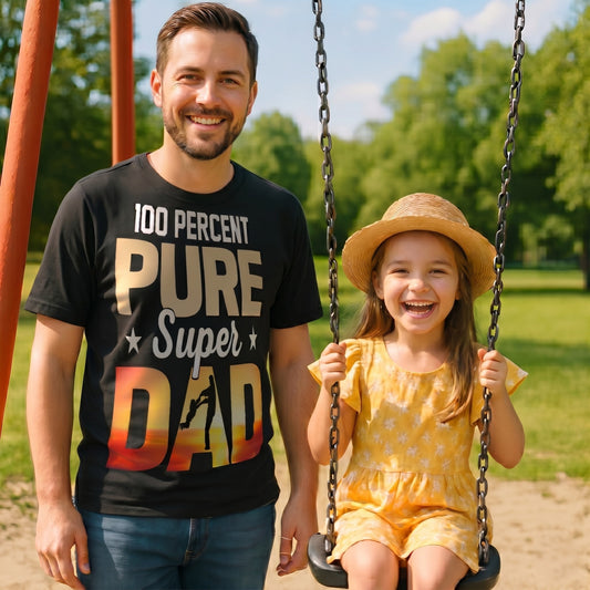 Man wearing a ‘100 Percent Pure Super Dad’ T‑shirt standing beside his child on a swing in a sunny park