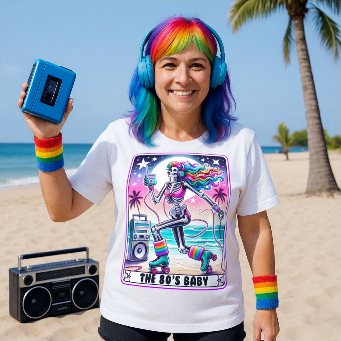 Woman wearing the 80s Baby retro graphic T-shirt featuring a colourful neon skeleton on roller skates with rainbow hair, headphones, cassette player, and boombox while standing on a tropical beach with vintage 80s accessories.