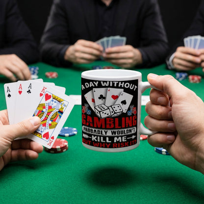 Person holding a funny gambling coffee mug featuring playing cards, poker chips, dice, and the slogan "A Day Without Gambling Probably Wouldn't Kill Me... But Why Risk It?" at a poker table.