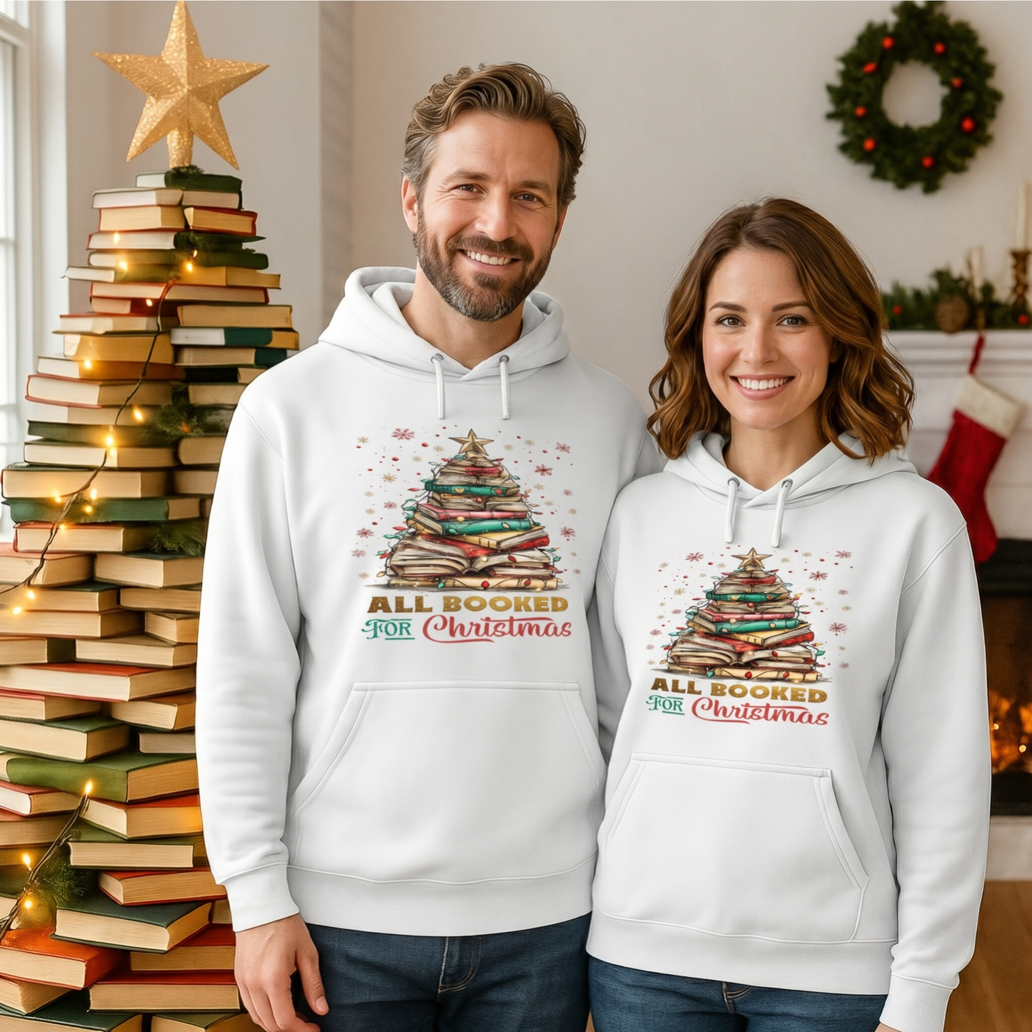 Couple wearing matching white All Booked For Christmas hoodies featuring a Christmas tree made from stacked books, standing beside a festive book Christmas tree decorated with fairy lights in a cosy holiday home.
