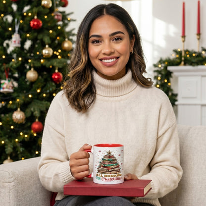 Woman in a cosy cream jumper holding the All Booked For Christmas mug with a festive book tree design, sitting beside a decorated Christmas tree with a red book on her lap in a warm holiday living room.