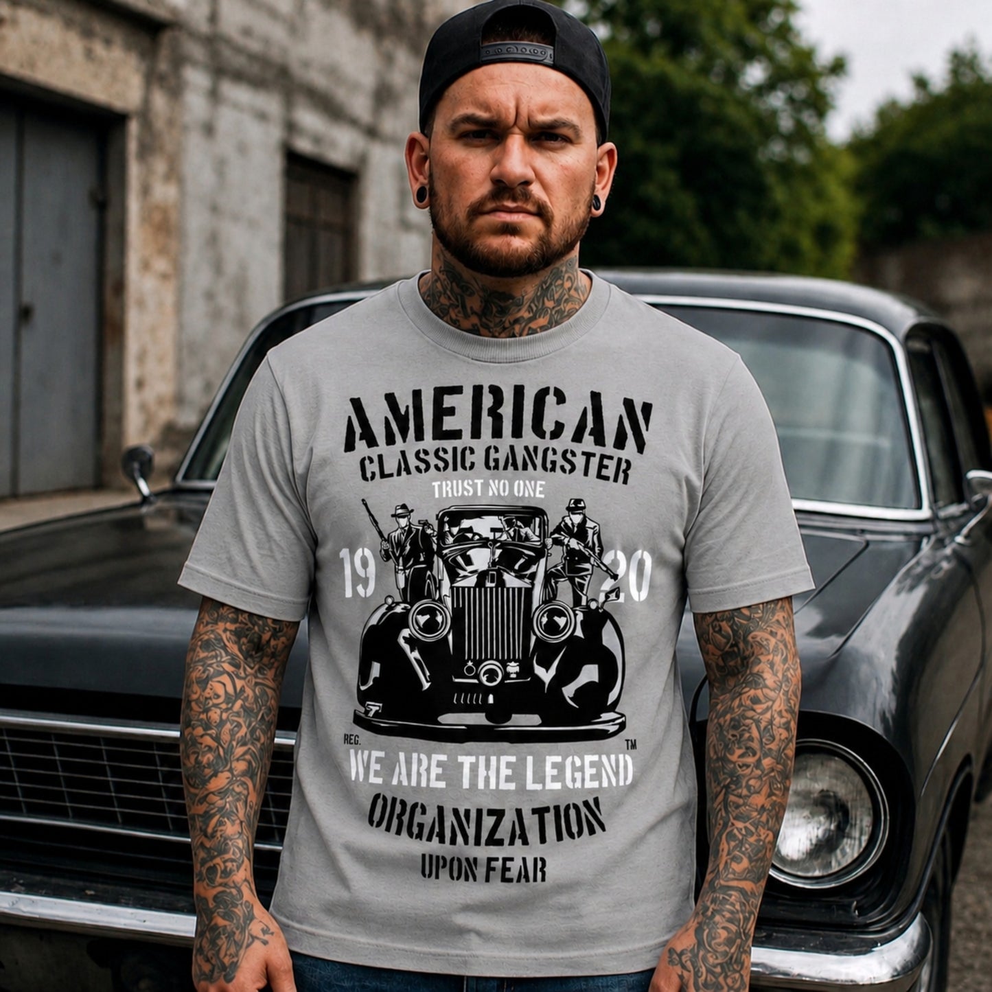 Lifestyle mockup of a person wearing the grey American Classic Gangster T-Shirt featuring a vintage automobile, gangster artwork, and distressed monochrome graphic while standing in front of a classic black car.