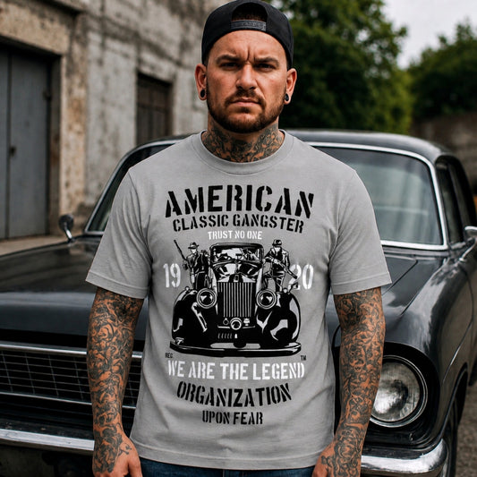 Lifestyle mockup of a person wearing the grey American Classic Gangster T-Shirt featuring a vintage automobile, gangster artwork, and distressed monochrome graphic while standing in front of a classic black car.