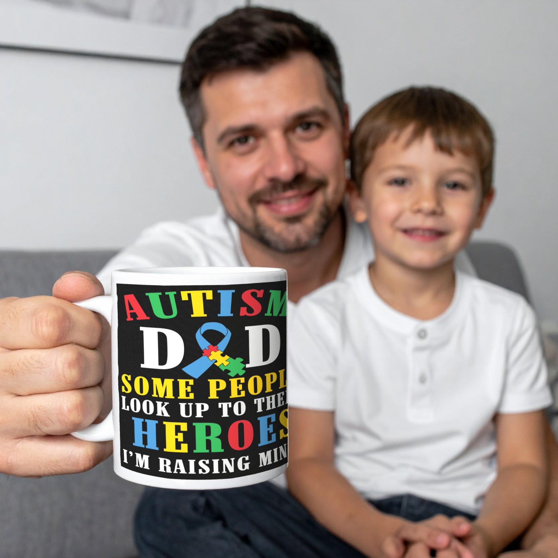 Man holding a white ceramic mug with colourful text that reads “Some people look up to their heroes, I’m raising mine” and features the autism awareness puzzle ribbon – perfect gift for autism dads