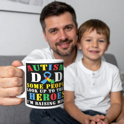 Man holding a white ceramic mug with colourful text that reads “Some people look up to their heroes, I’m raising mine” and features the autism awareness puzzle ribbon – perfect gift for autism dads