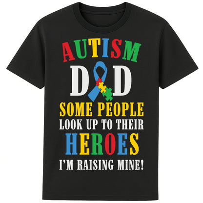 A black t-shirt with bold, colorful text reading: AUTISM DAD SOME PEOPLE LOOK UP TO THEIR HEROES I'M RAISING MINE," The design celebrates fatherhood and pride in raising a child with autism.