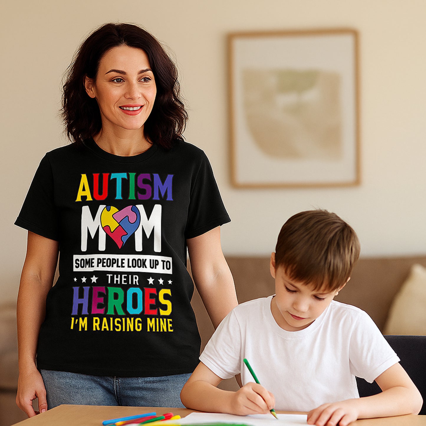 A woman wearing a black t-shirt with bold, colourful text reading: "AUTISM MOM SOME PEOPLE LOOK UP TO THEIR HEROES I'M RAISING MINE," The design celebrates motherhood and pride in raising a child with autism.
