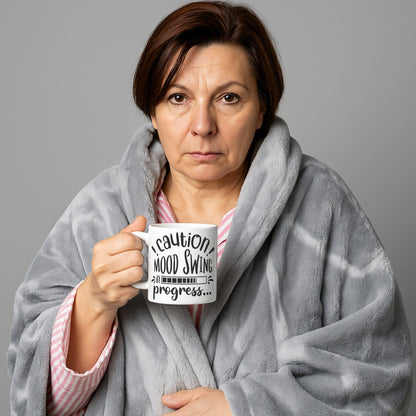Woman wrapped in a blanket holding a mug with text that reads “Caution! Mood Swing in Progress…” — a humorous gift idea, especially relatable for menopause mood swings.