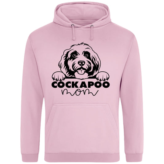 Pink hoodie with a playful design featuring a cartoon Cockapoo dog peeking over the bold text 'Cockapoo Mom' in a mix of black and pink fonts. The dog has a cute expression with its tongue out and paws hanging over the text."