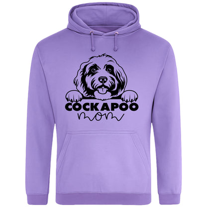 A lavender hoodie with a playful design featuring a cartoon Cockapoo dog peeking over the bold text 'Cockapoo Mom' in a mix of black and pink fonts. The dog has a cute expression with its tongue out and paws hanging over the text."