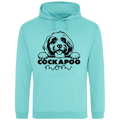 A peppermint hoodie with a playful design featuring a cartoon Cockapoo dog peeking over the bold text 'Cockapoo Mom' in a mix of black and pink fonts. The dog has a cute expression with its tongue out and paws hanging over the text."