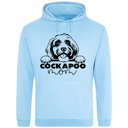 A Sky Blue hoodie with a playful design featuring a cartoon Cockapoo dog peeking over the bold text 'Cockapoo Mom' in a mix of black and pink fonts. The dog has a cute expression with its tongue out and paws hanging over the text."