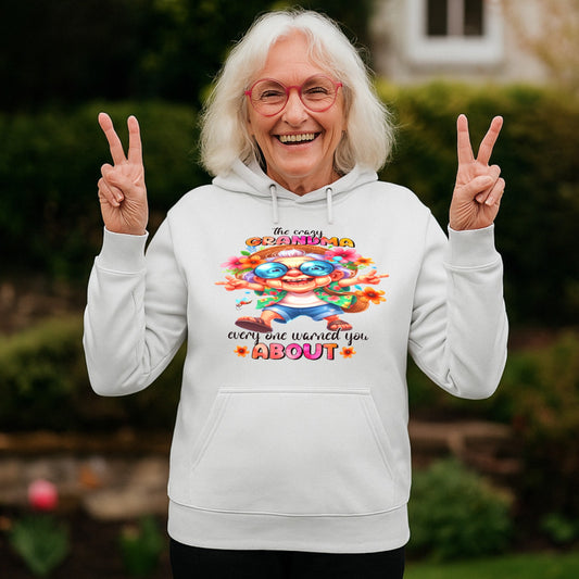 Smiling grandmother wearing a white Crazy Grandma Everyone Warned You About hoodie while making peace signs in a garden, funny grandma hoodie, gift for grandma, grandmother birthday gift, and Grandparents Day gift.