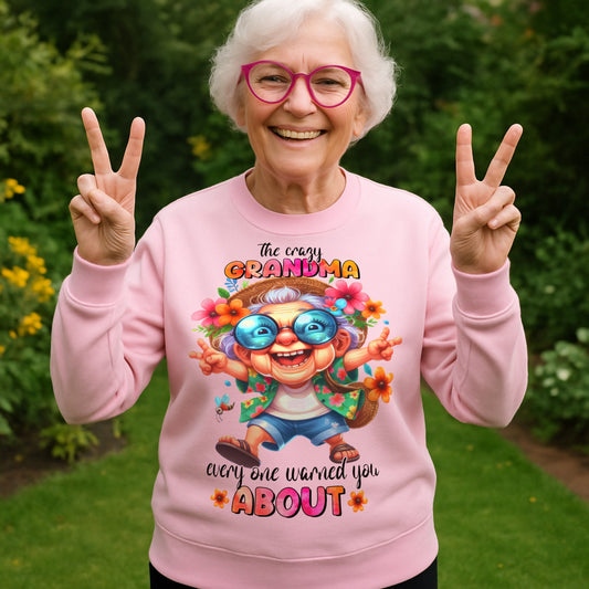 Smiling grandmother wearing a pink Crazy Grandma Everyone Warned You About sweatshirt while making peace signs in a garden, funny grandma sweatshirt, grandmother gift, birthday gift, and Grandparents Day gift.