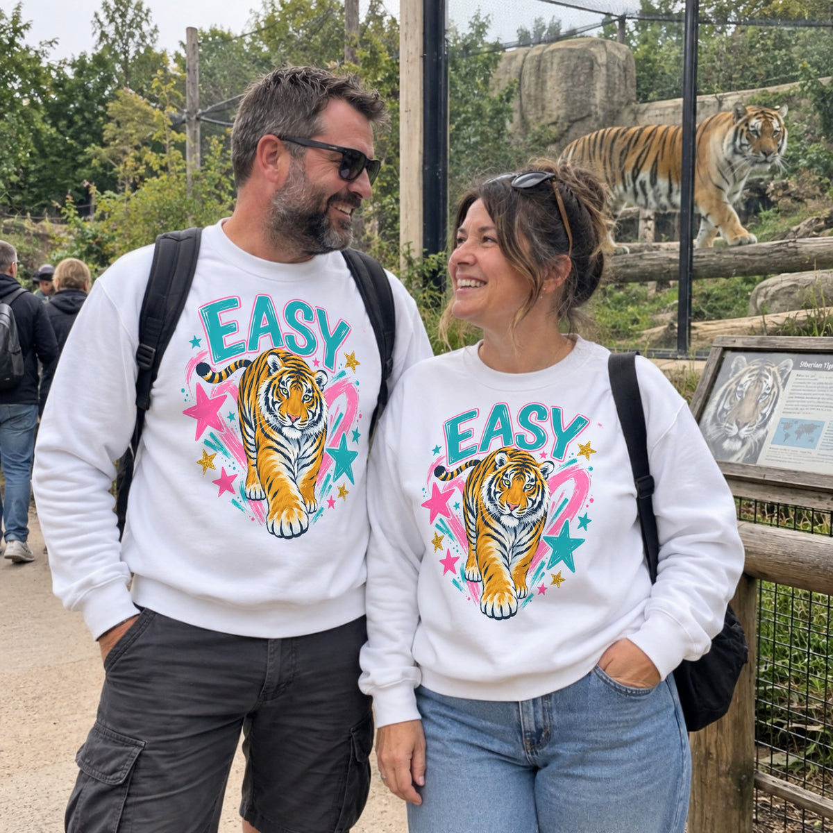 Man and woman wearing matching Easy Tiger graphic sweatshirts beside a tiger enclosure at a wildlife park.