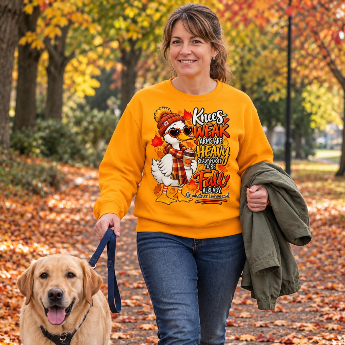 Woman walking her dog in autumn wearing a funny fall goose sweatshirt with colourful Knees Weak Arms Are Heavy seasonal artwork.