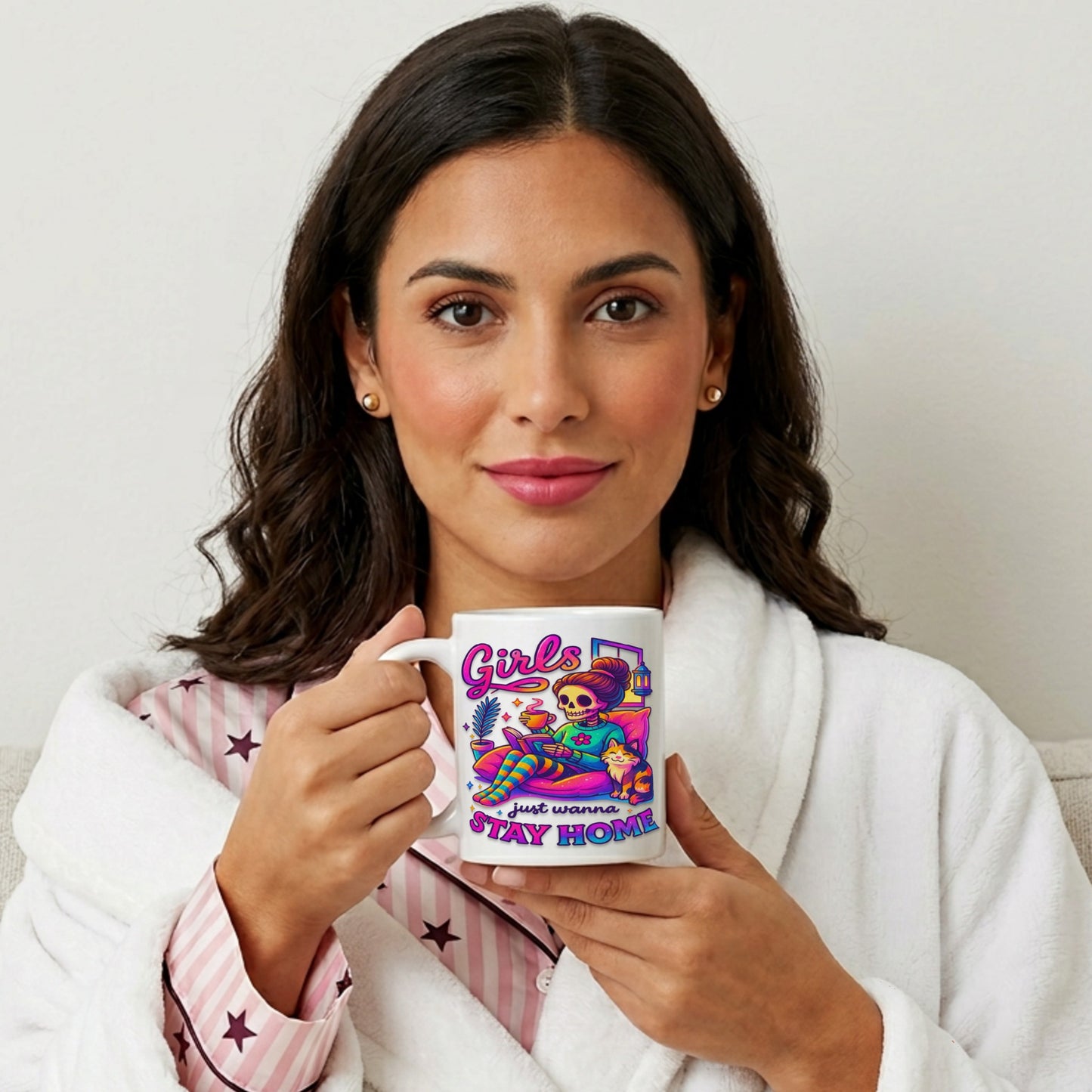 Woman in cosy pyjamas and a fluffy dressing gown holding the Girls Just Wanna Stay Home Mug featuring colourful skeleton artwork, a funny homebody coffee mug for introverts, book lovers, and cat lovers.