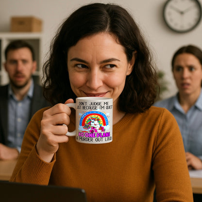Woman holding a  White ceramic mug with a rainbow unicorn holding an axe, framed by a rainbow and clouds, with humorous text about being quiet and planning a murder.