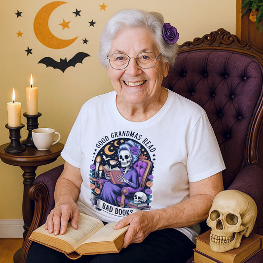 Smiling grandma wearing the Good Grandmas Read Bad Books T-Shirt while reading a book in a cosy chair with candles, skull décor, and gothic accents, funny grandma t shirt for book lovers and spooky readers.