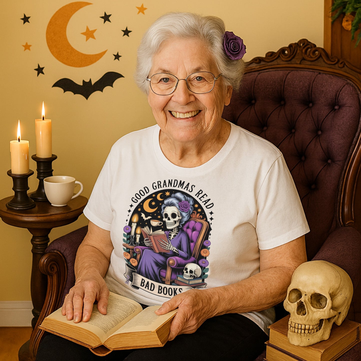 Woman sitting in a chair holding a book, wearing a t-shirt with a graphic design, “Good Grandmas Read Bad Books T-Shirt  in a room decorated with Halloween elements.