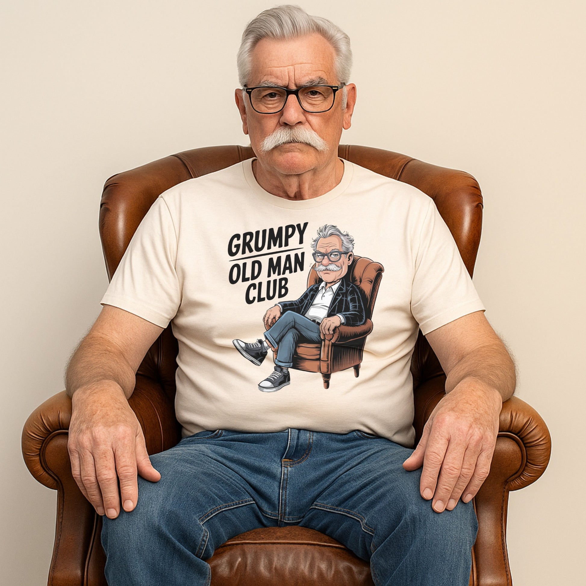 An older man wearing a "Grumpy Old Man Club" t-shirt with a cartoon of a grumpy man in an armchair, sitting seriously in a leather chair.