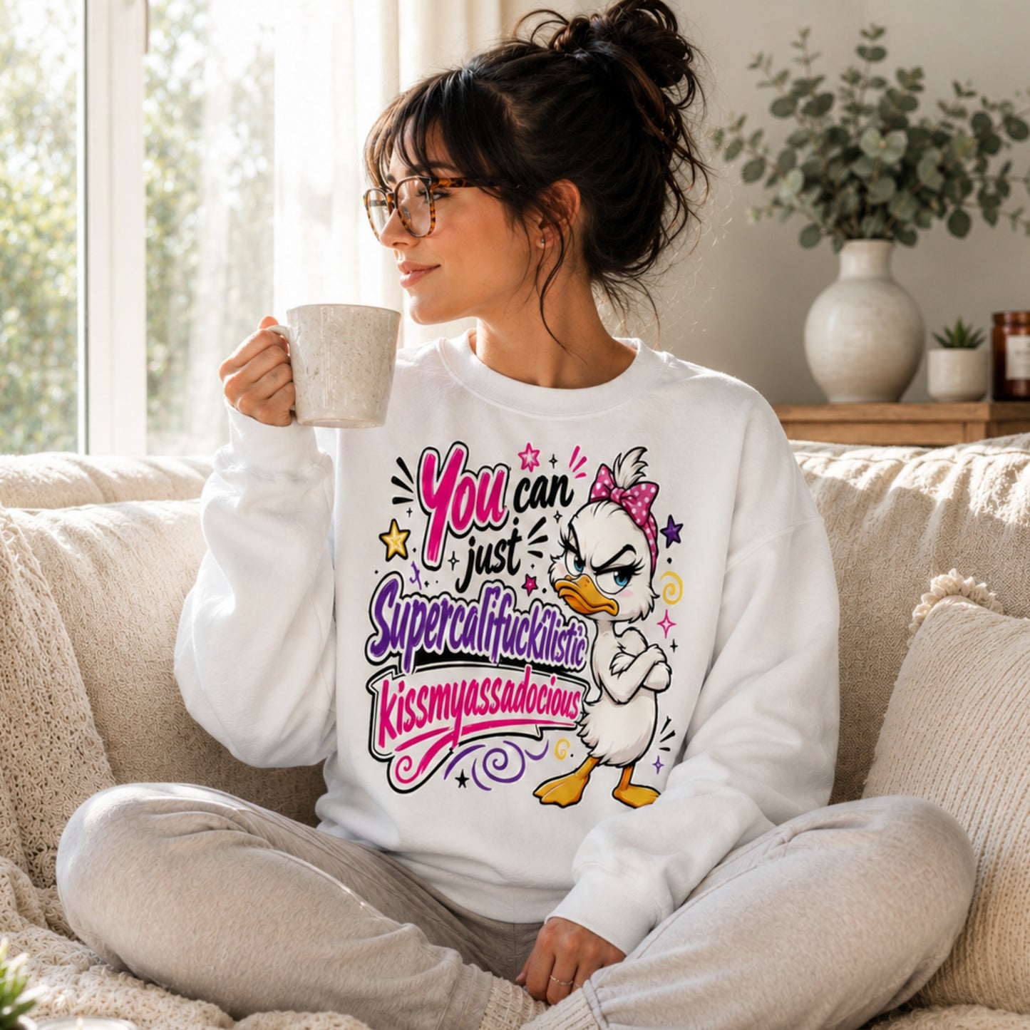 Woman with dark hair in a messy bun and glasses wearing a white Grumpy Sarcastic Duck sweatshirt while relaxing on a cream sofa with a coffee in a bright sunlit living room, cosy unisex funny graphic sweatshirt.