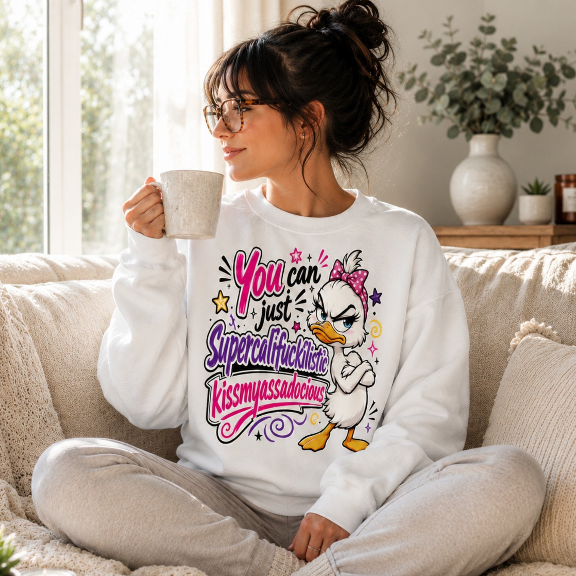 Woman with dark hair in a messy bun and glasses wearing a white Grumpy Sarcastic Duck sweatshirt while relaxing on a cream sofa with a coffee in a bright sunlit living room, cosy unisex funny graphic sweatshirt.