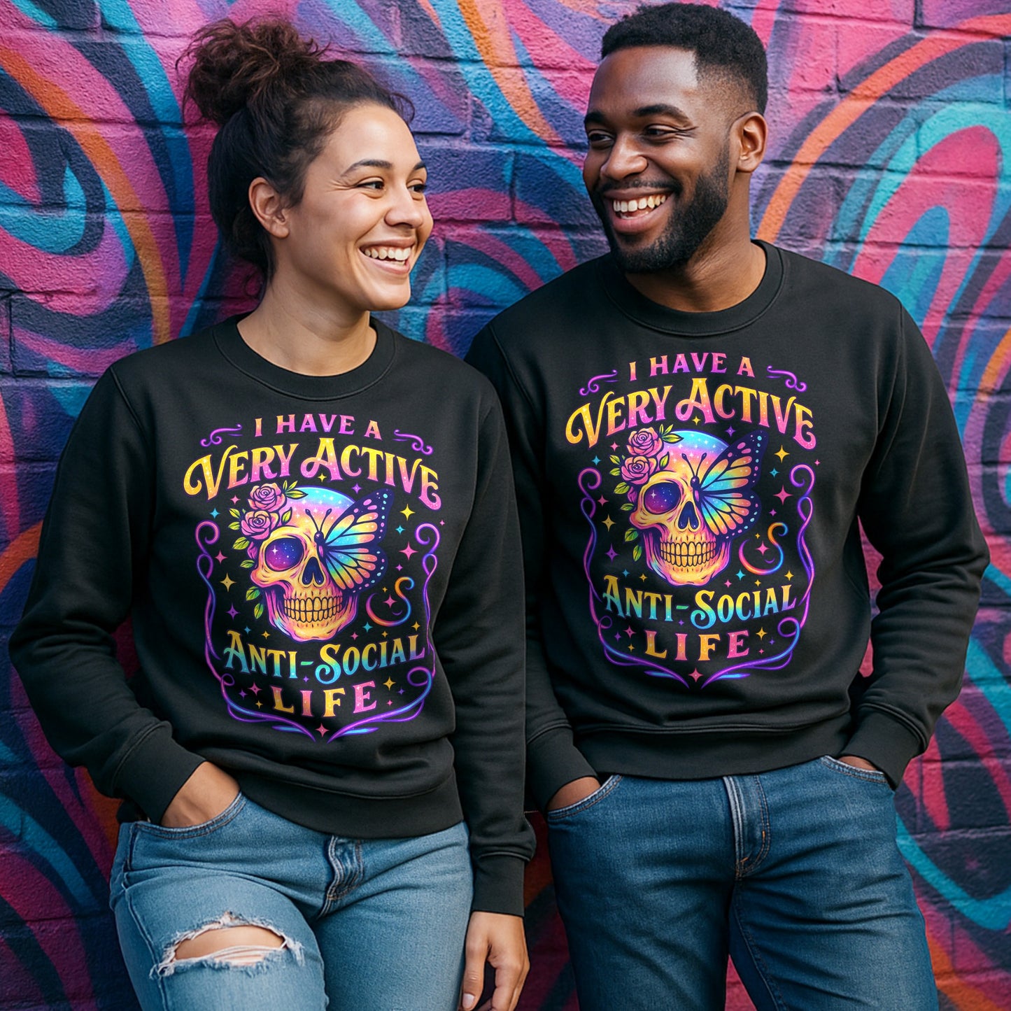Male and female models wearing the I Have A Very Active Anti Social Life sweatshirt featuring a colourful neon sugar skull, butterfly, and floral graphic on black unisex graphic sweatshirts against a vibrant graffiti wall.