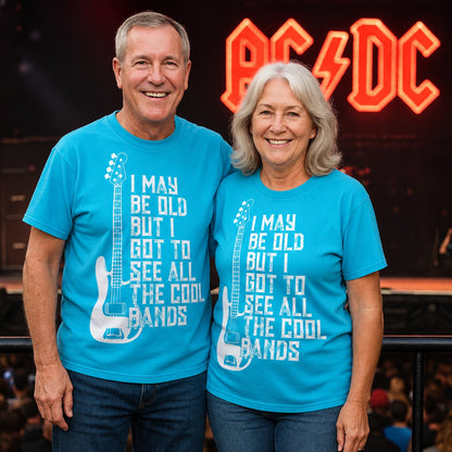 Older man and woman wearing matching blue "I May Be Old But I Got To See All The Cool Bands" T-shirts with a distressed guitar graphic, standing at a live rock concert venue with a stage and illuminated band sign in the background.