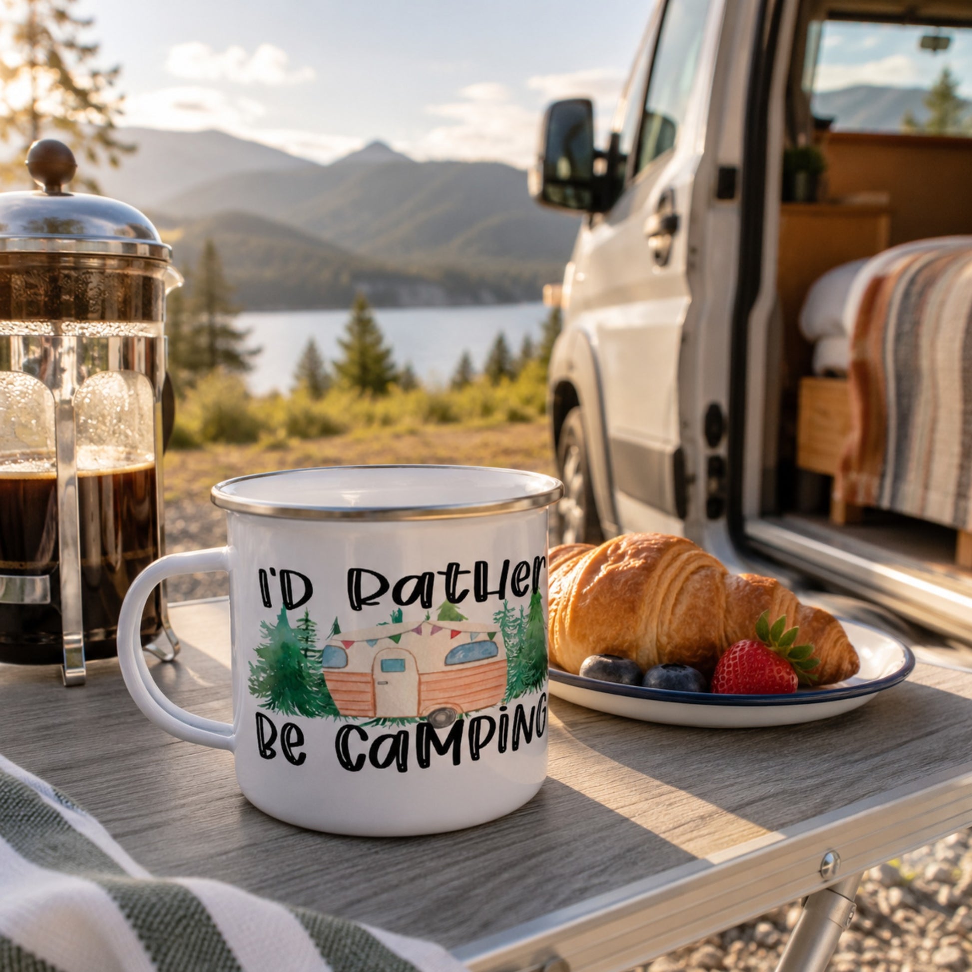 Outdoor enamel camping mug with an "I'd Rather Be Camping" vintage camper design displayed on a breakfast table beside a campervan, French press coffee, and pastries overlooking a scenic mountain lake campsite at sunrise.