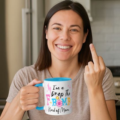 Smiling woman holding an "I'm A Drop The F-Bomb Kind Of Mom" funny mom coffee mug with blue handle and colourful retro lettering while making a middle finger gesture in a kitchen.