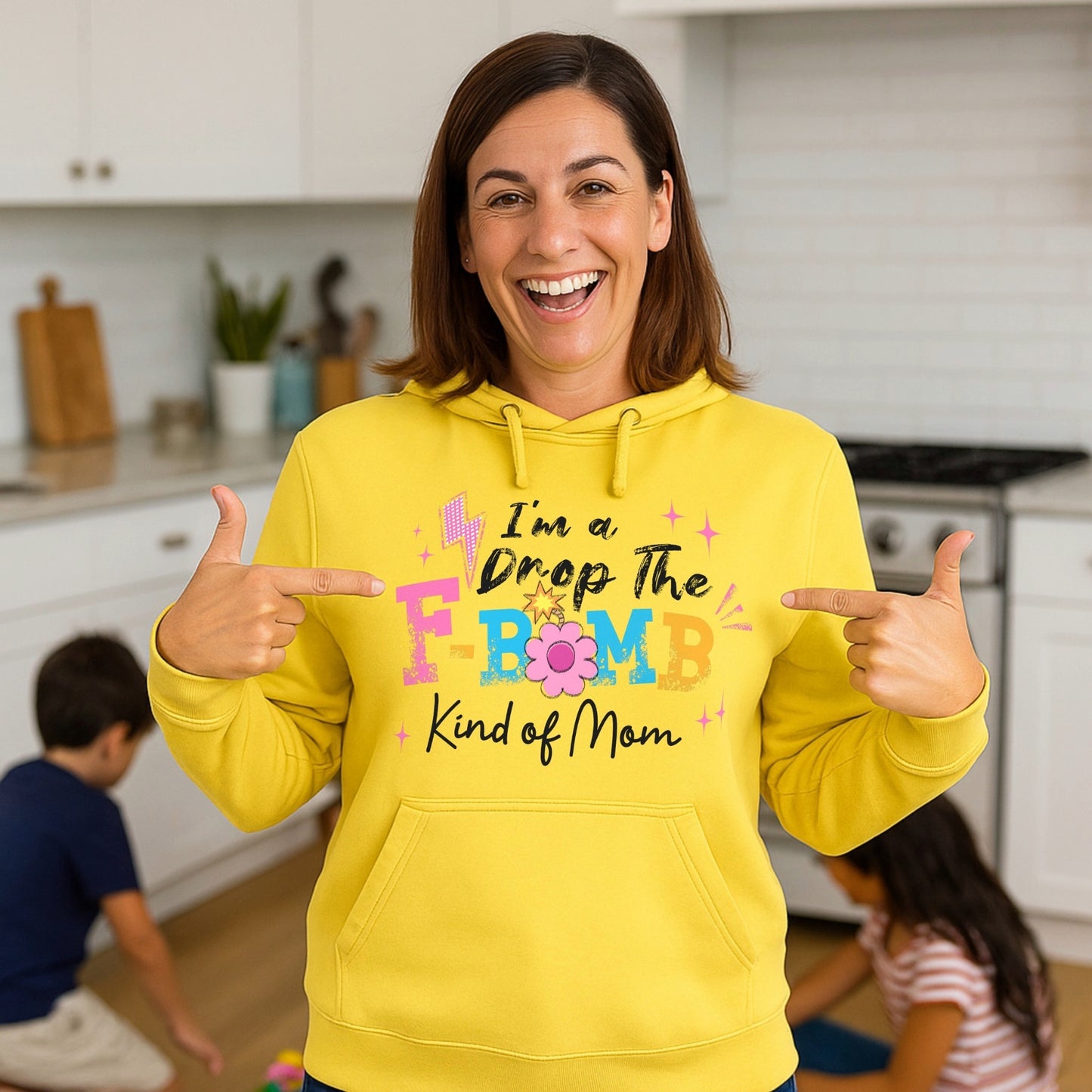 Smiling woman wearing a yellow "I'm A Drop The F-Bomb Kind Of Mom" hoodie with colourful retro lettering while pointing at the design in a kitchen with children in the background.