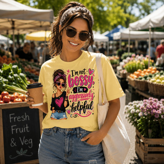 Woman wearing a yellow "I'm Not Bossy, I'm Aggressively Helpful" graphic T-shirt at an outdoor farmers market, relaxed fit unisex tee with longer sleeves, holding a takeaway coffee and canvas tote bag.