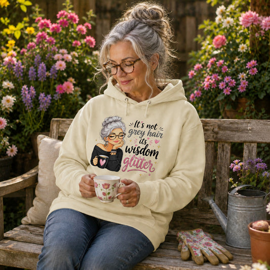 An older woman with grey hair in a bun sits on a wooden garden bench holding a mug, wearing a vanilla milkshake colored hoodie with a graphic of an elderly woman and the text 'It's not grey hair, it's wisdom glitter'.
