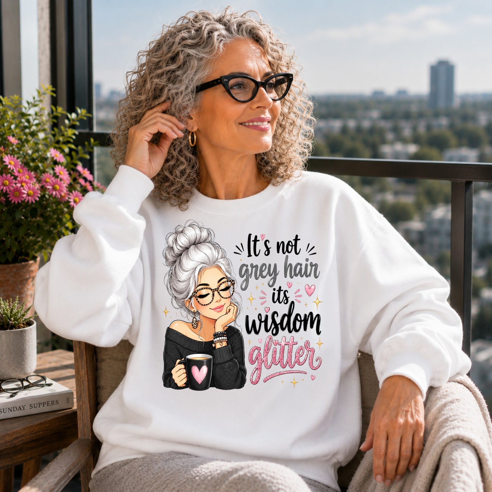 Woman with naturally curly grey hair wearing a white It's Not Grey Hair It's Wisdom Glitter sweatshirt while relaxing on a sunny balcony with a city view.