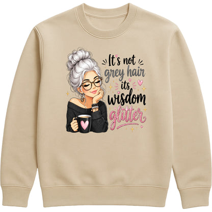 A nude colour crewneck sweatshirt featuring a graphic print of an elderly woman with grey hair, glasses, and a black top holding a mug with a pink heart, next to the text 'It's not grey hair, it's wisdom glitter' in black and pink lettering.