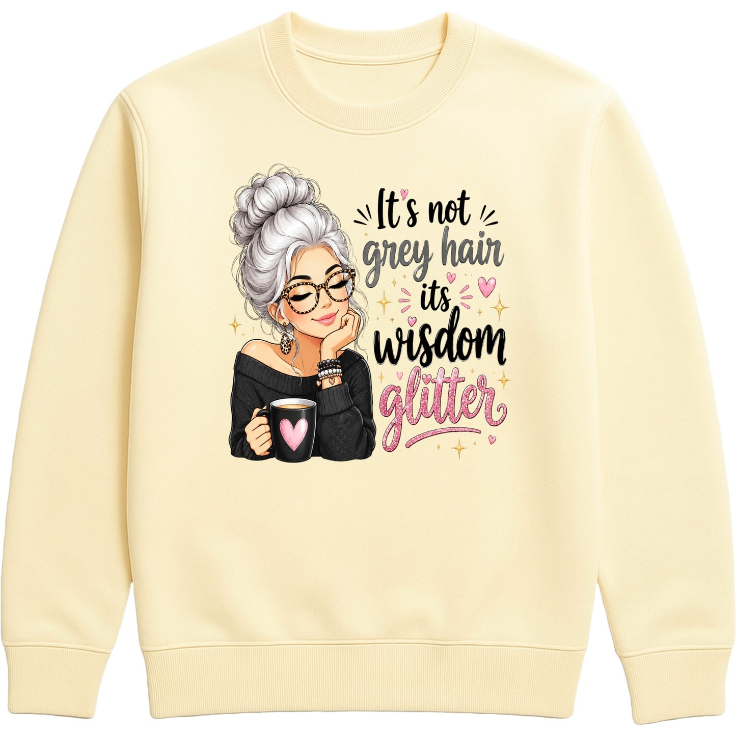 A vanilla milkshake colored crewneck sweatshirt featuring a graphic of an elderly woman with grey hair, glasses, and a black top, holding a mug with a pink heart. To her right is the text 'It's not grey hair it's wisdom glitter' in black and pink lettering with small pink hearts and gold star accents.