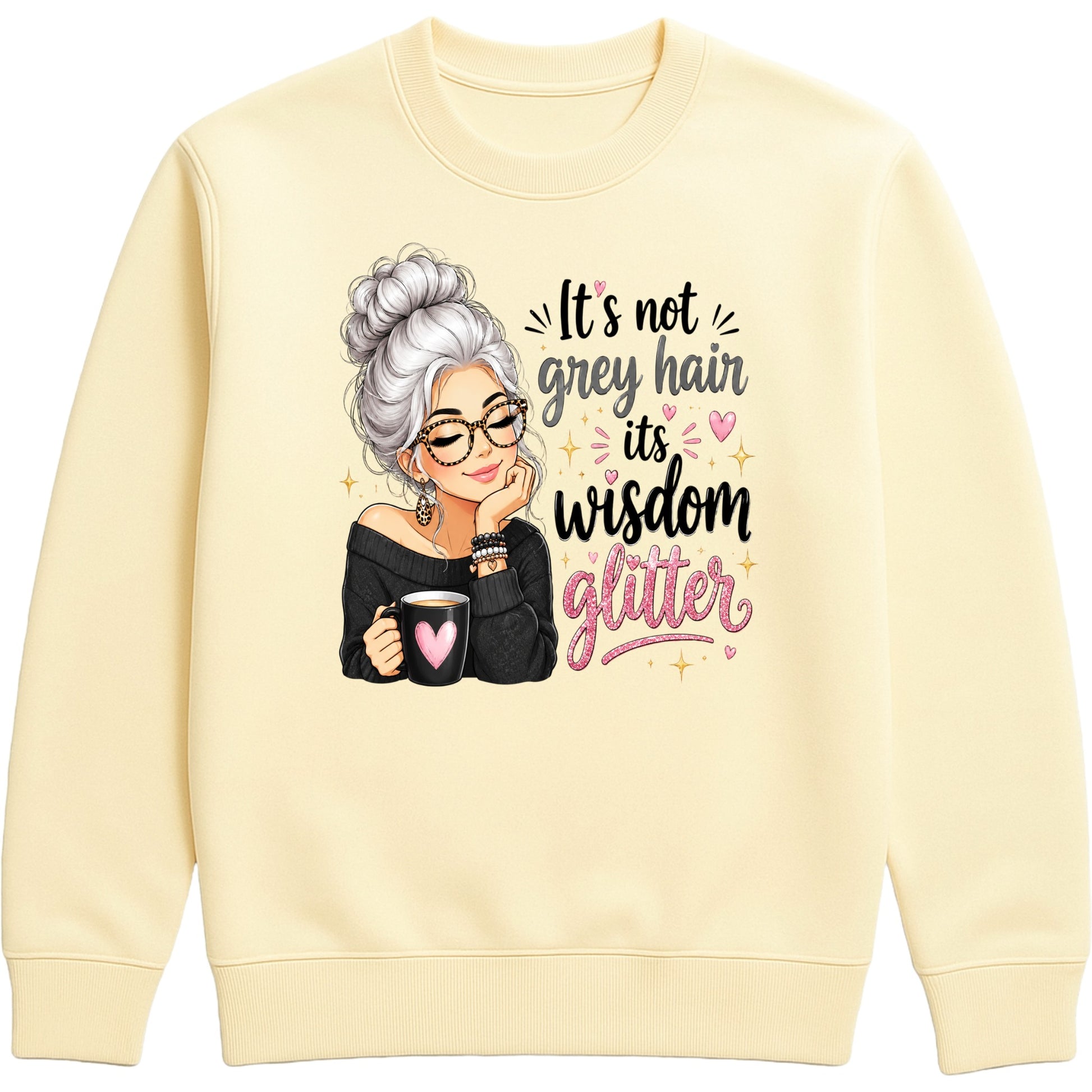 A vanilla milkshake colored crewneck sweatshirt featuring a graphic of an elderly woman with grey hair, glasses, and a black top, holding a mug with a pink heart. To her right is the text 'It's not grey hair it's wisdom glitter' in black and pink lettering with small pink hearts and gold star accents.