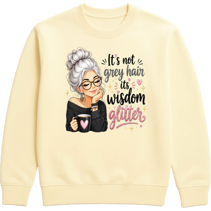 A vanilla milkshake colored crewneck sweatshirt featuring a graphic of an elderly woman with grey hair, glasses, and a black top, holding a mug with a pink heart. To her right is the text 'It's not grey hair it's wisdom glitter' in black and pink lettering with small pink hearts and gold star accents.