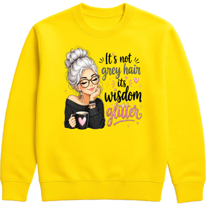 A bright yellow crewneck sweatshirt featuring a graphic of an older woman with grey hair, glasses, and a black top holding a mug, next to the text 'It's not grey hair its wisdom glitter' in black and pink lettering.