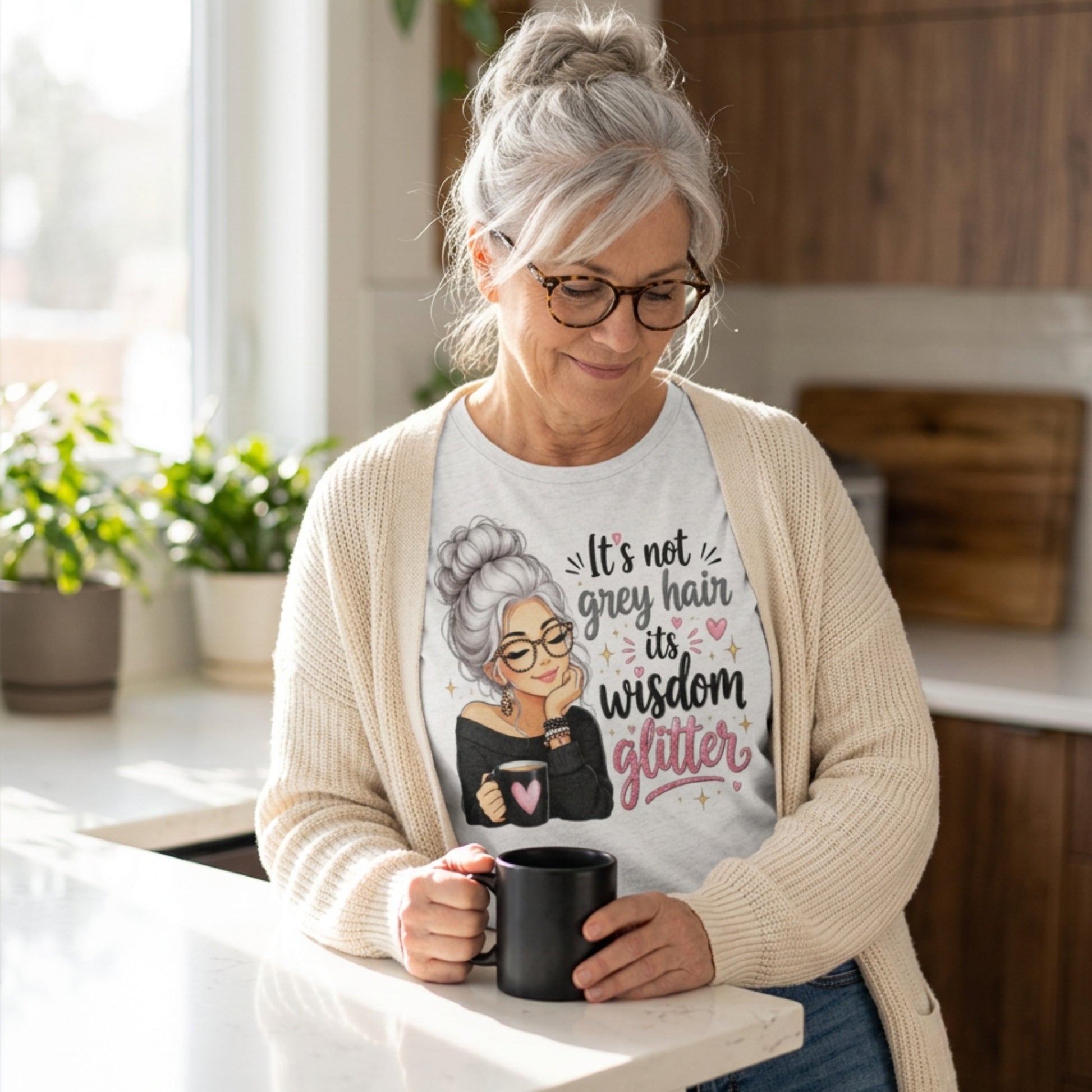 An older woman with grey hair and glasses stands in a bright kitchen, wearing a cream cardigan over a white graphic t-shirt. The t-shirt features an illustration of an older woman with grey hair and glasses, with the text 'It's not her grey hair, it's her wisdom glitter' in black and pink lettering.