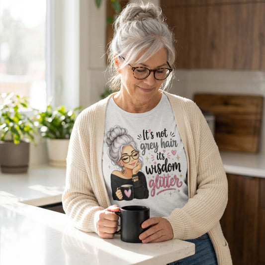 An older woman with grey hair and glasses stands in a bright kitchen, wearing a cream cardigan over a white graphic t-shirt. The t-shirt features an illustration of an older woman with grey hair and glasses, with the text 'It's not her grey hair, it's her wisdom glitter' in black and pink lettering.
