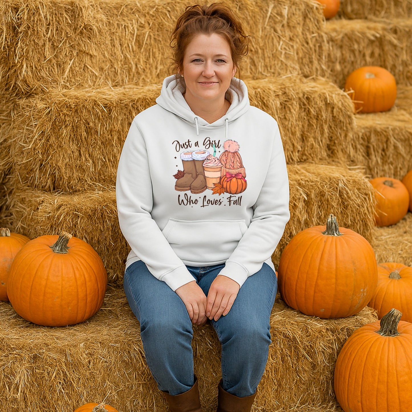 Woman wearing the Just a Girl Who Loves Fall hoodie while sitting beside pumpkins and hay bales at a pumpkin patch, featuring cosy autumn boots, a pumpkin spice latte, knitted beanie and pumpkin artwork.