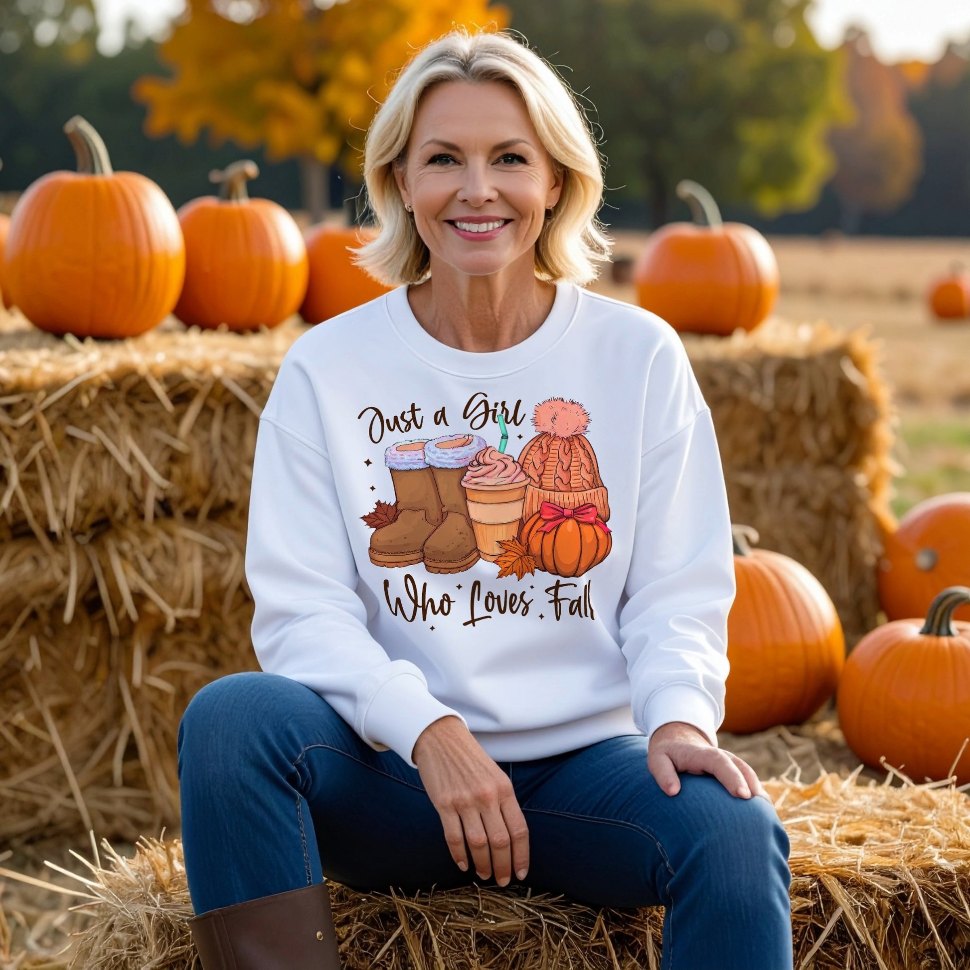 Woman wearing a white sweatshirt with fall-themed design sitting on hay bales with pumpkins in the background.